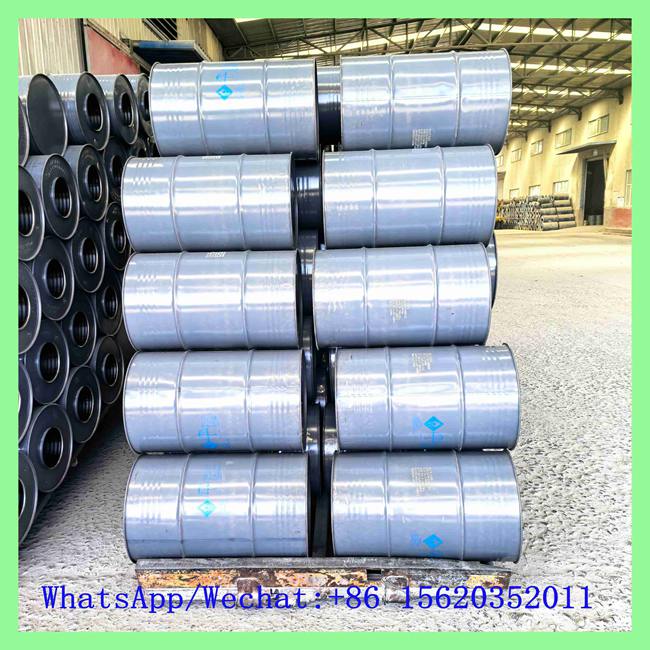 Grey metal chemical drums with blue hazardous material labels neatly horizontally stacked on wooden pallets in a factory warehouse setting.