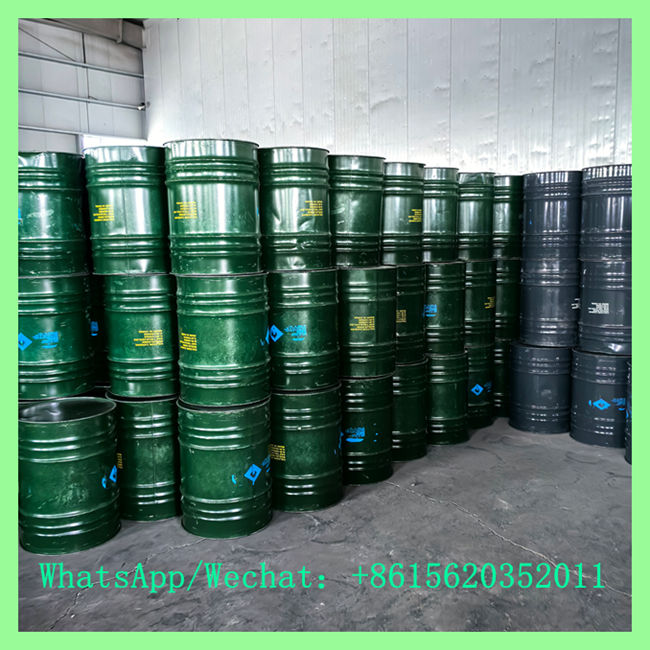 Green metal industrial drums stacked neatly in a warehouse, featuring blue hazard diamond stickers and yellow text labels.