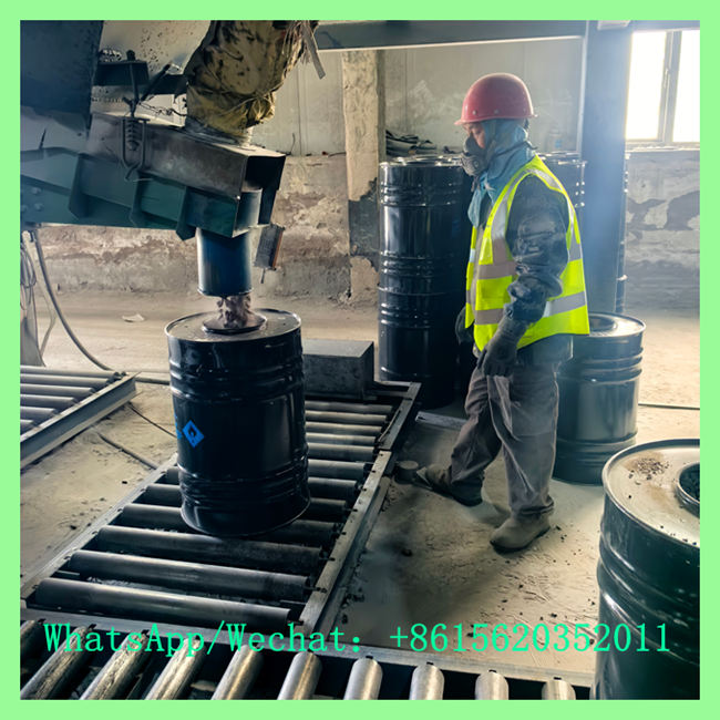 Factory worker wearing a hard hat and safety vest guiding a black chemical drum along a roller conveyor during the filling process.