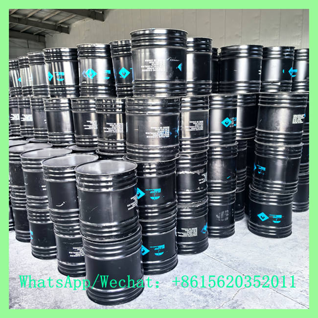 Multiple black industrial metal drums stacked in organized rows inside a storage warehouse, showing blue hazard symbols.