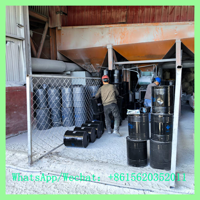 Workers in protective gear moving and stacking black metal drums near factory processing hoppers and equipment.
