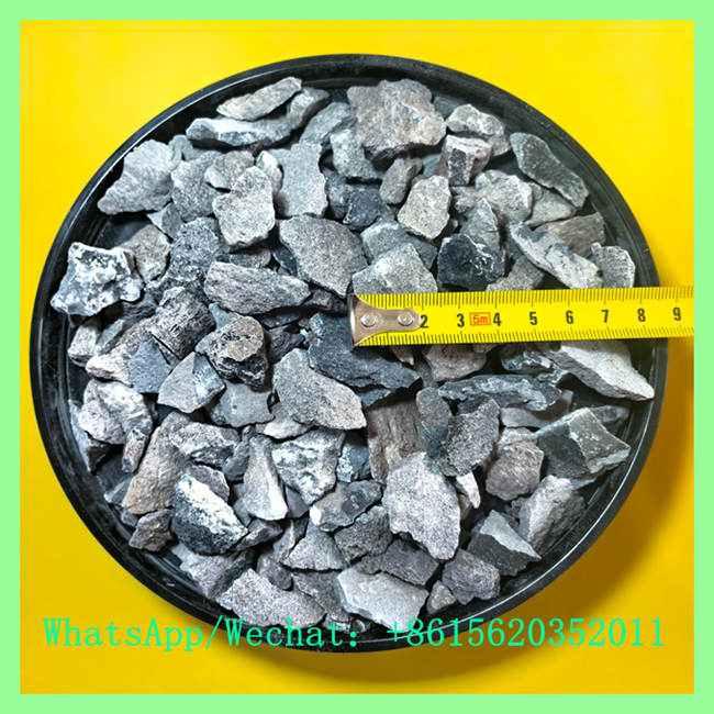 Top-down view of dark grey crushed calcium carbide lumps inside a black tray with a yellow measuring tape showing particle size.